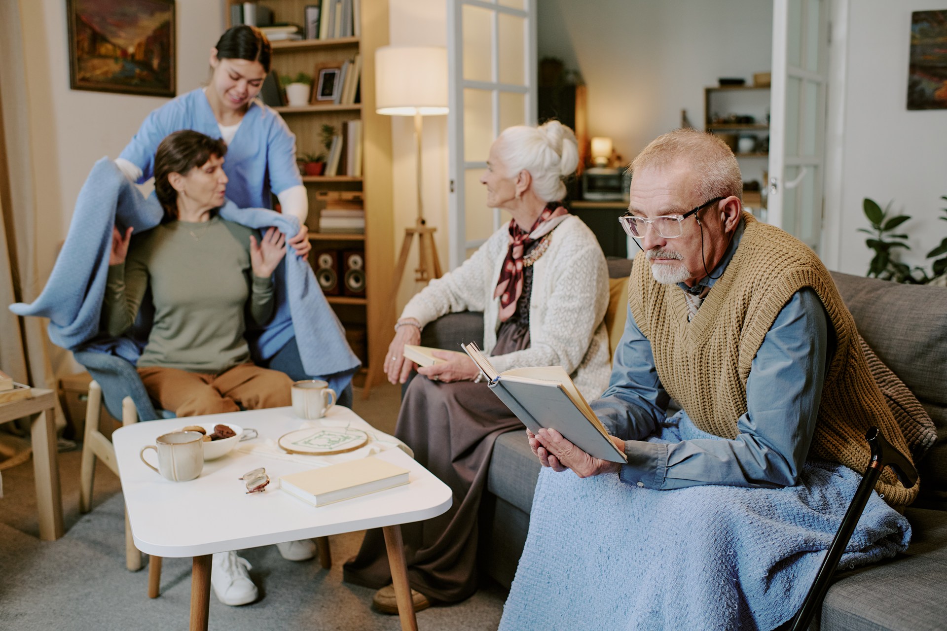 Seniorzy odpoczywają w salonie, a opiekunka okrywa kobietę kocem w prywatnym domu opieki w Gdańsku
